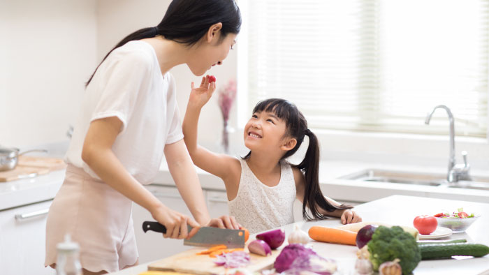 Young girl feeding her mother a grape tomato while they chop vegetables in the kitchen