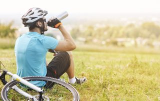 A man in bike riding gear sits on the grass and drinks water from a water bottle next to his bicycle