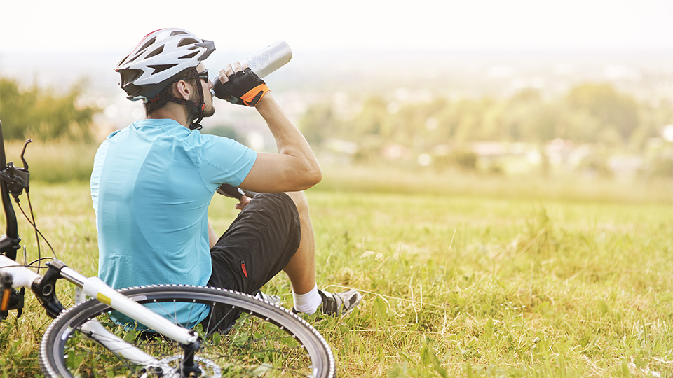 A man in bike riding gear sits on the grass and drinks water from a water bottle next to his bicycle