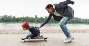 A dad pushing his kid on a skateboard by the water