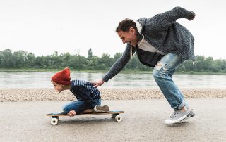 A dad pushing his kid on a skateboard by the water