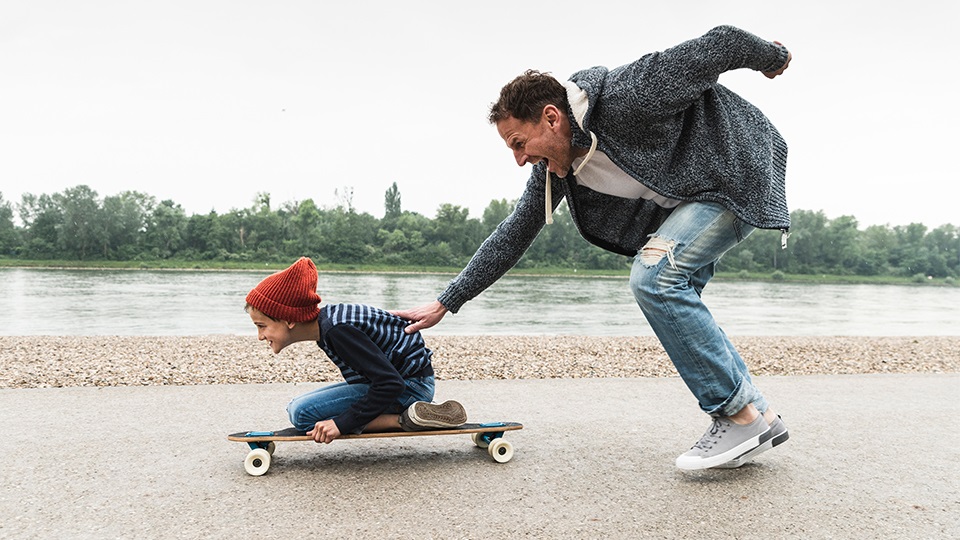 A dad pushing his kid on a skateboard by the water