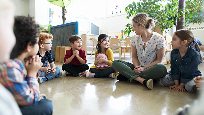 Children sitting in a circle on the floor with a teacher. One girl is holding a pink stuffed animal