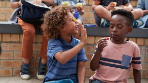 Boy with a grape against his closed eye reaching out for a high-five from another boy holding a piece of fruit