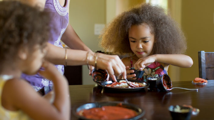 Two young girls putting cheese and sauce on their pizza at the kitchen table