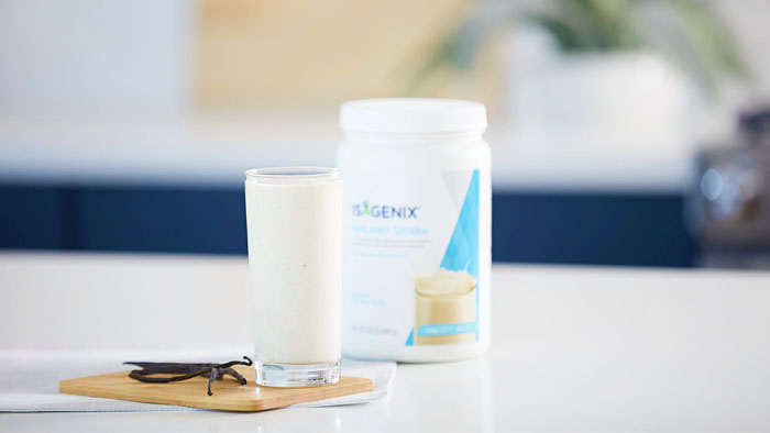 Canister of IsaLean Shake and blended IsaLean Shake in a glass on a wood board with vanilla.