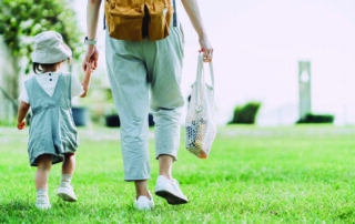 Parent holding hands with their child while walking on the grass and carrying a reusable bag