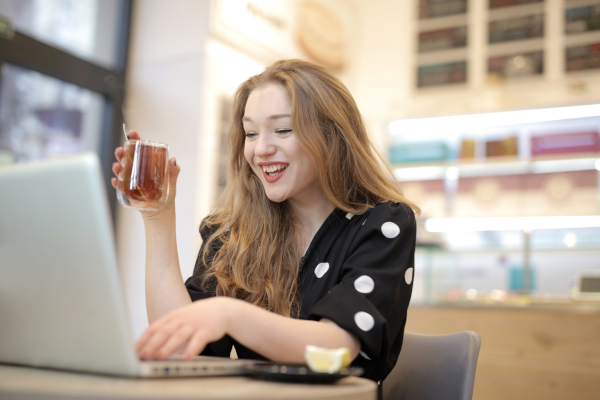 Redheaded woman holding a clear mug of tea while working on a laptop