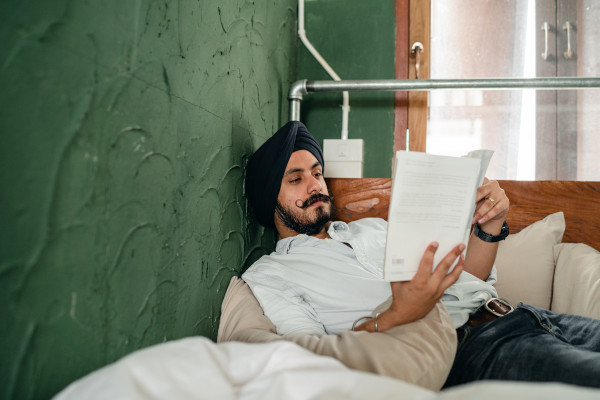 Man with a mustache reclining on a bed and reading a book alone